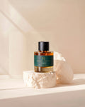 A bottle of French Parfums OMD Diffused Oil of Amber fragrance, featuring a dark green label and black cap, displayed on a textured white stone pedestal against a warm beige background.